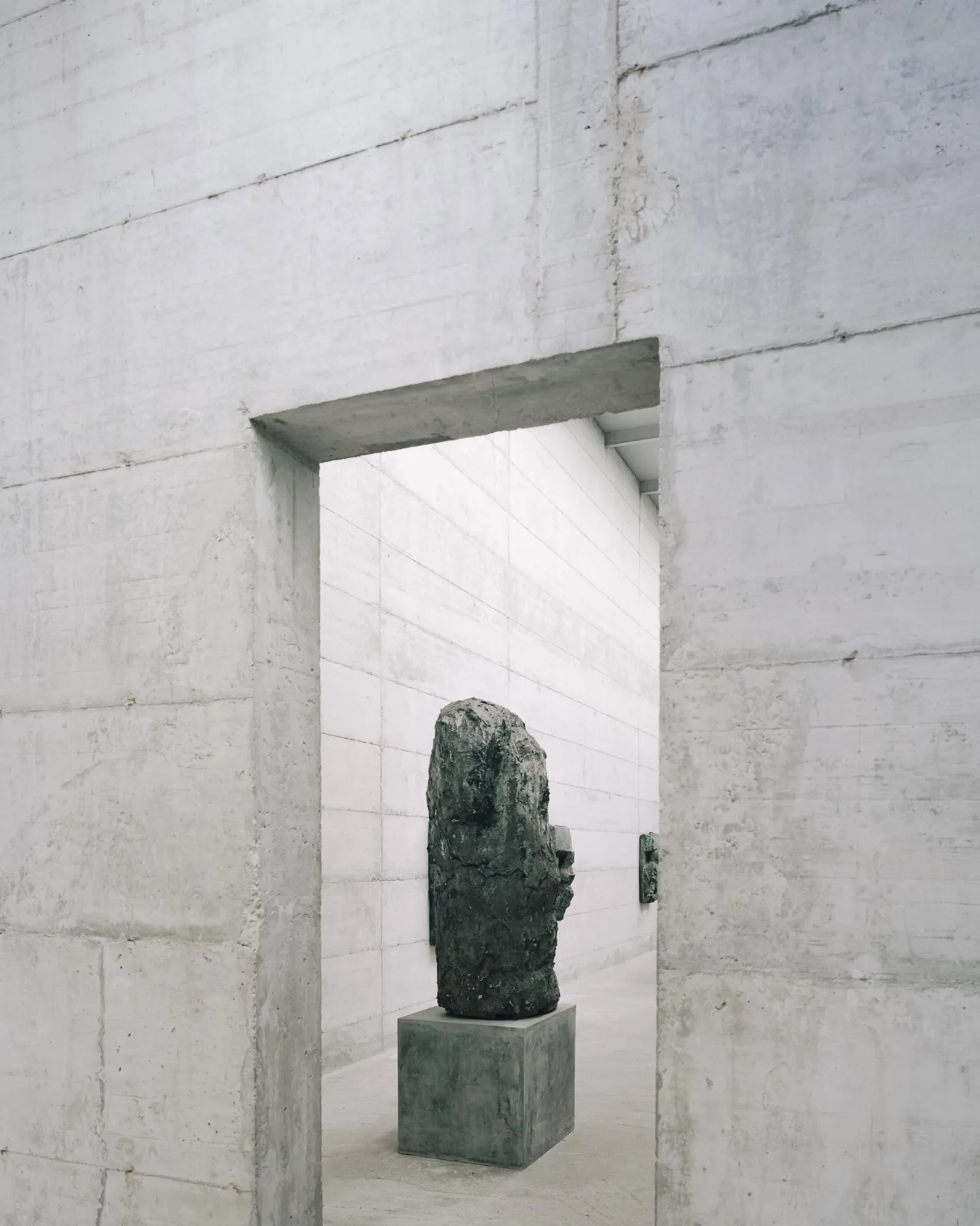 La Congiunta Museum by Peter Märkli — A dark stone sculptural head mounted on a concrete plinth occupies the center of a narrow gallery corridor, framed by the rectangular aperture of a concrete...