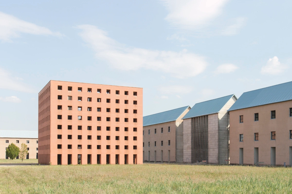 San Cataldo Cemetery by Aldo Rossi — A terracotta brick cubic volume dominates the foreground, its facade pierced by a regular grid of square voids that create rhythmic visual repetition across...
