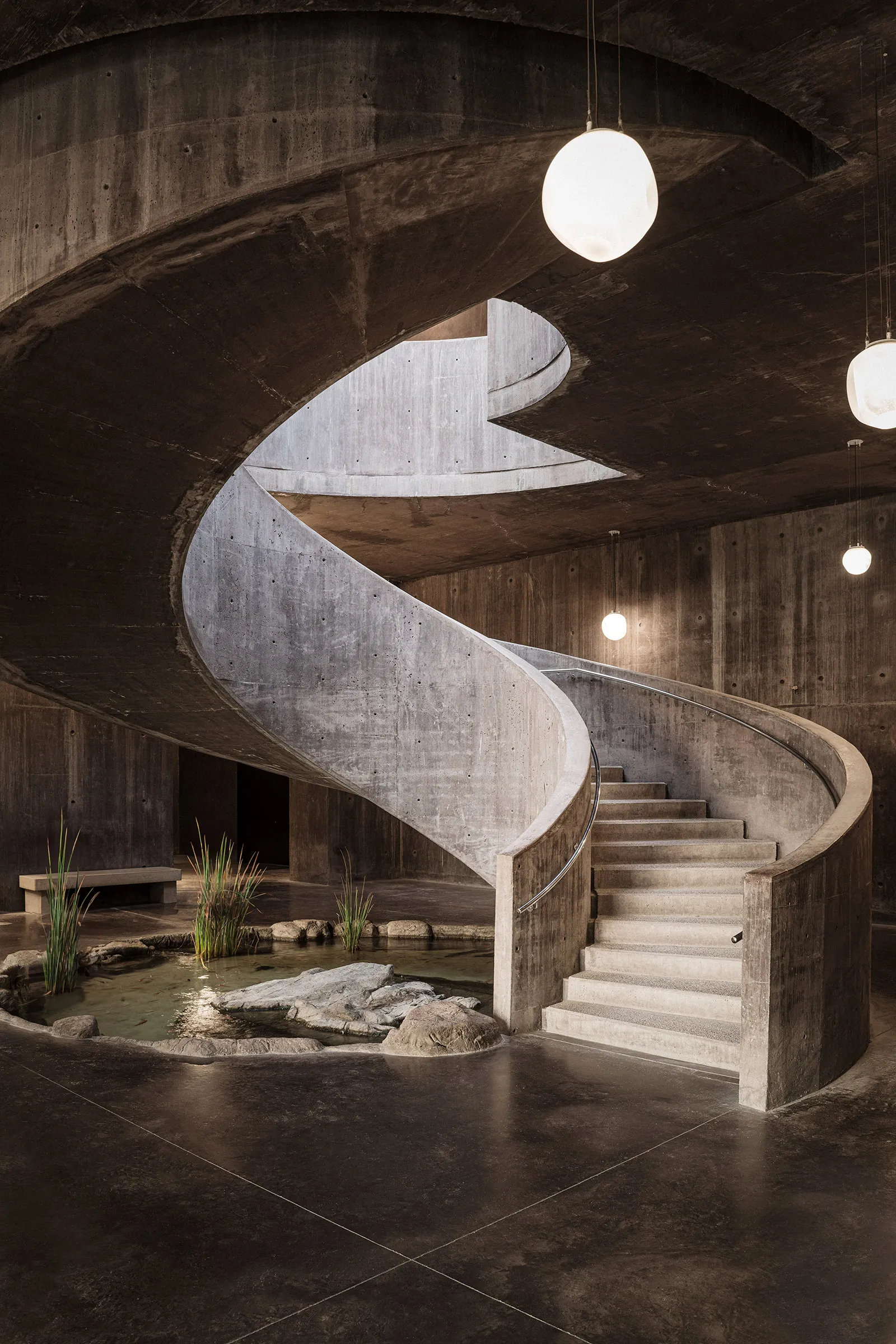 Sea of Cortez Research Center by Tatiana Bilbao — A monolithic concrete spiral staircase descends through an underground chamber, its sculptural form illuminated by suspended spherical pendant lights.