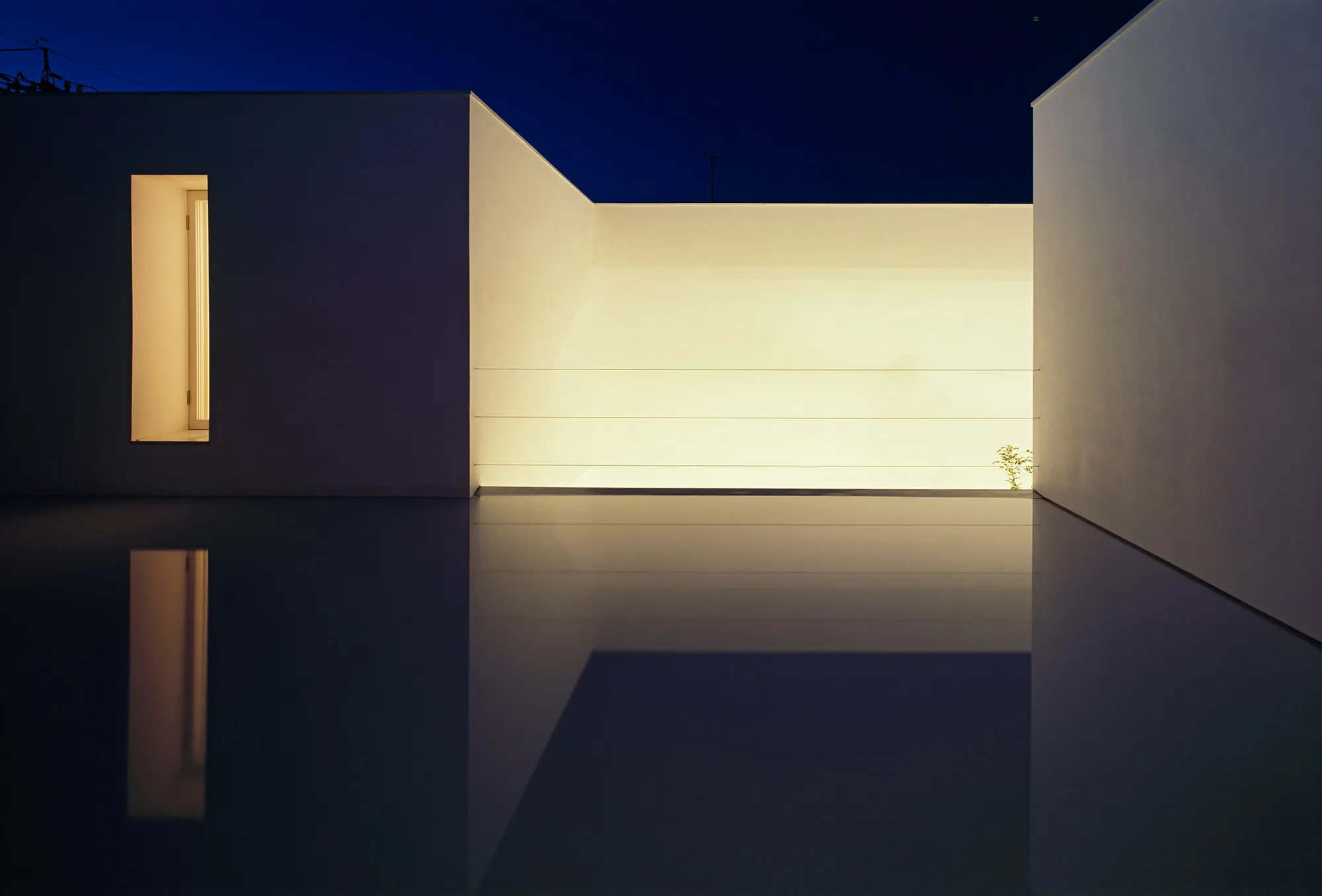 White Cave House by Takuro Yamamoto — Night photograph of White Cave House's recessed courtyard void, illuminated from within by warm artificial light that contrasts sharply with the deep blue...