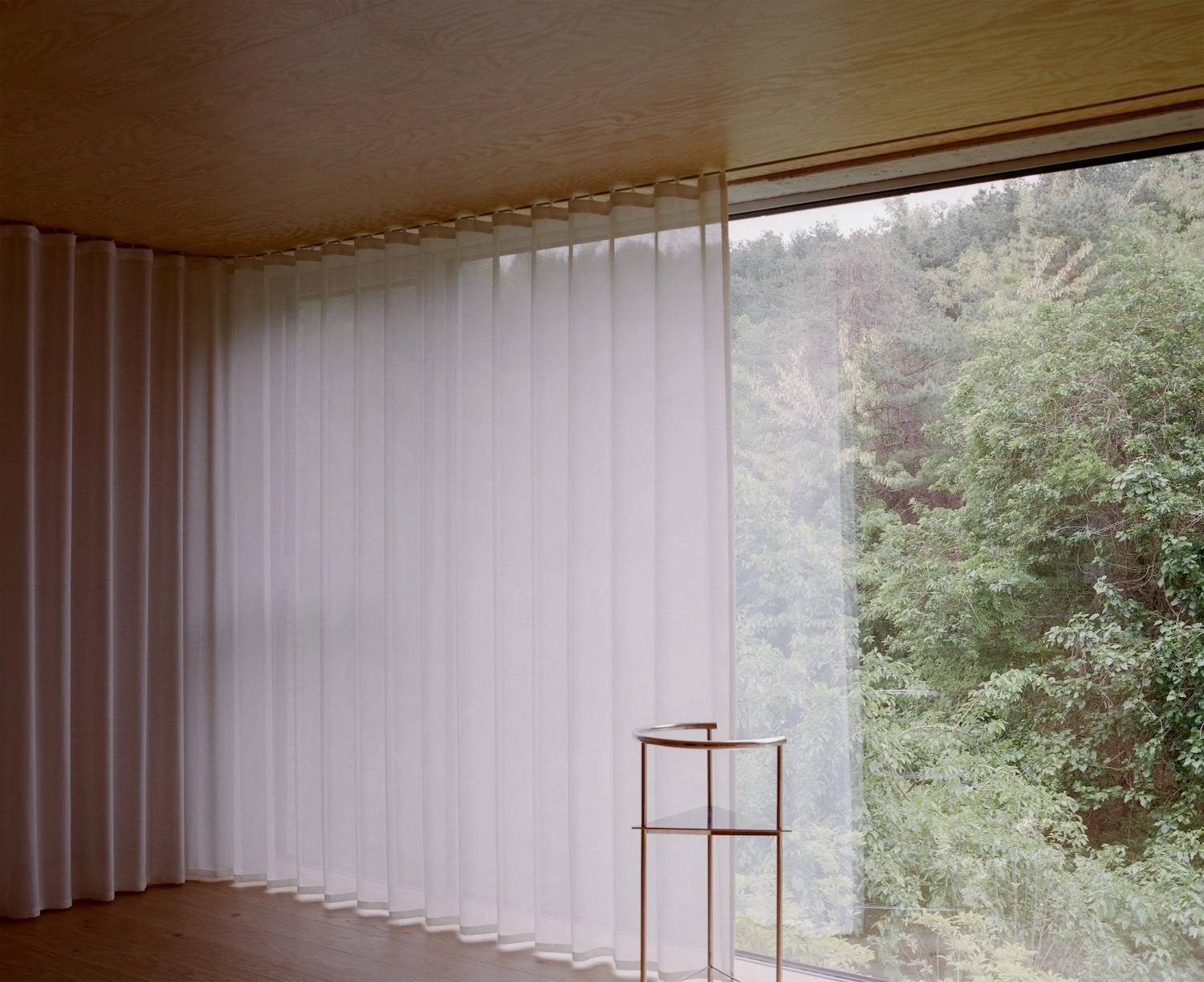 Raw House by Order Matter — A reading nook positioned within the Raw House's expansive southern glazing, where sheer linen curtains filter views of the wooded hillside beyond.