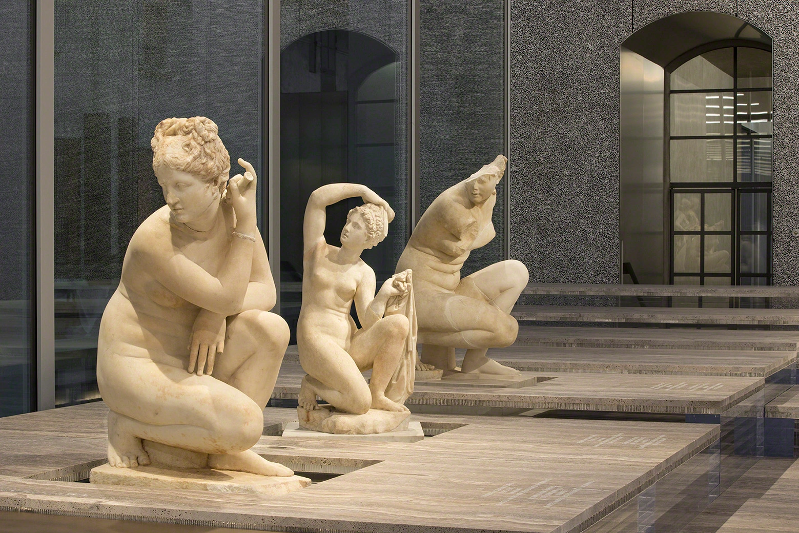 Fondazione Prada by OMA — Three neoclassical marble sculptures occupy a wooden deck within Fondazione Prada's courtyard, positioned before a light grey textured aggregate facade with...