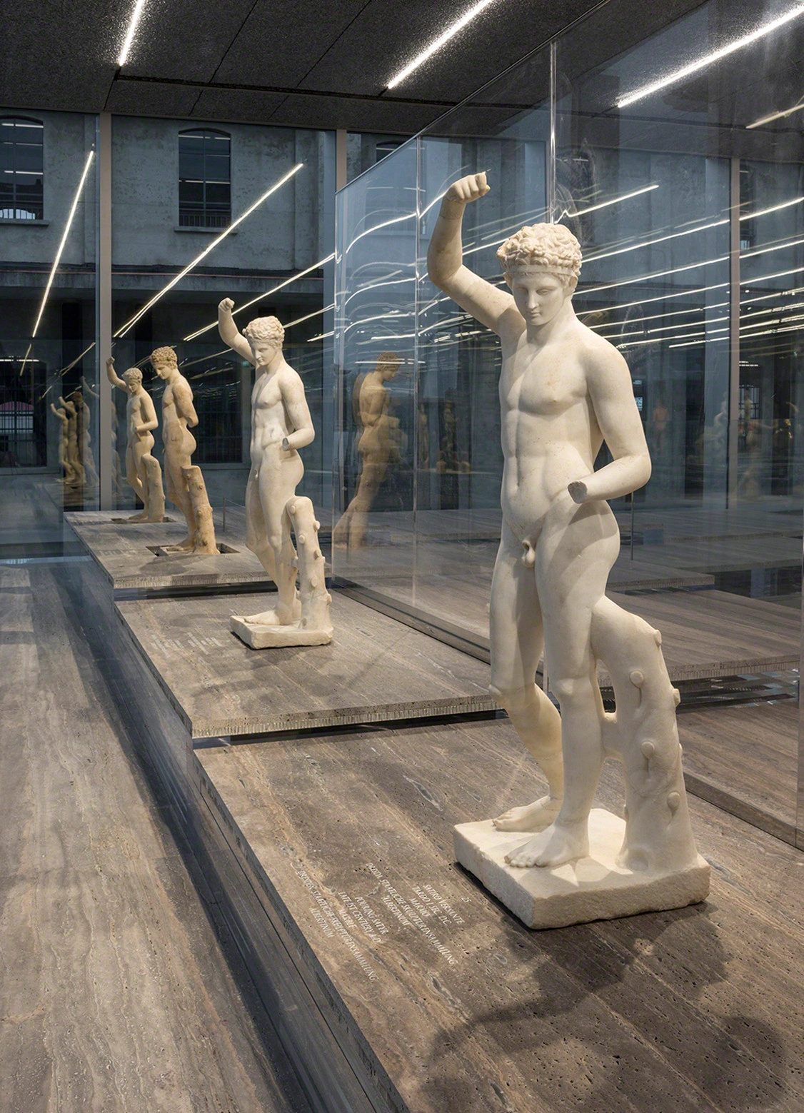 Fondazione Prada by OMA — A row of classical plaster cast sculptures—primarily torsos and full figures in white and cream tones—displayed in a glass-enclosed corridor.