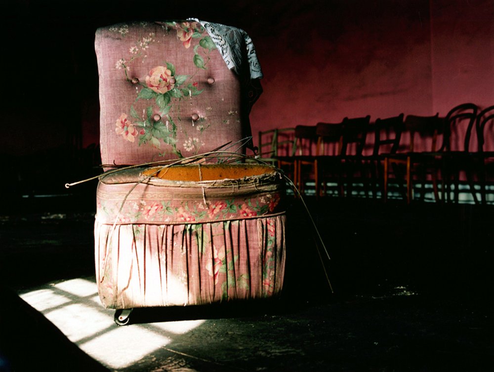 After Magdalene by Ethna Rose O'Regan — A stuffed upholstered chair sits isolated in harsh institutional light, its rose-patterned fabric faded and worn.