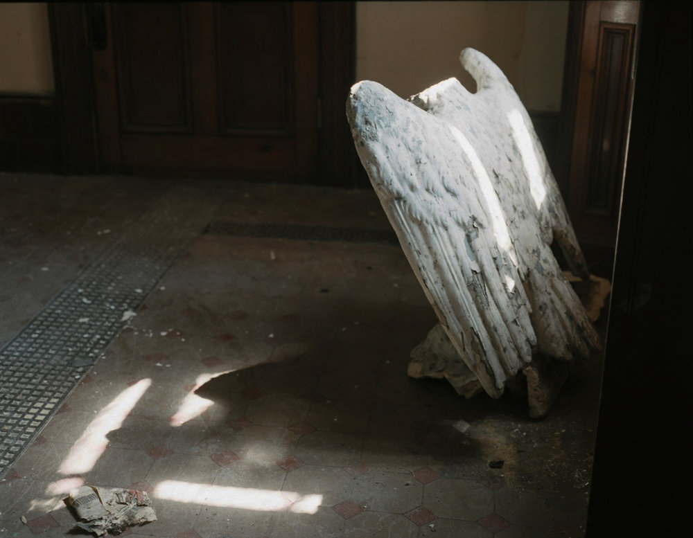 After Magdalene by Ethna Rose O'Regan — A weathered plaster cast of wings leans against an industrial concrete floor in harsh diagonal sunlight.