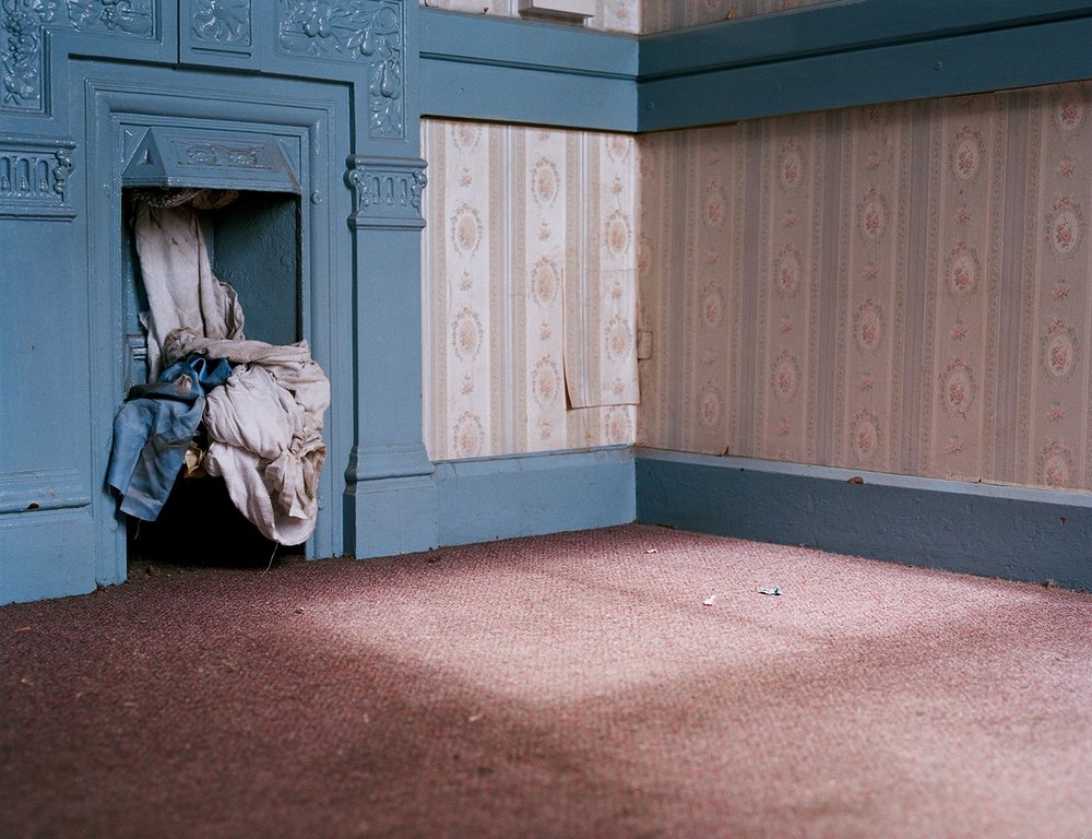 After Magdalene by Ethna Rose O'Regan — A corner of an abandoned institutional dormitory, its blue-painted wooden door frame fitted with decorative relief molding contrasts sharply against striped...