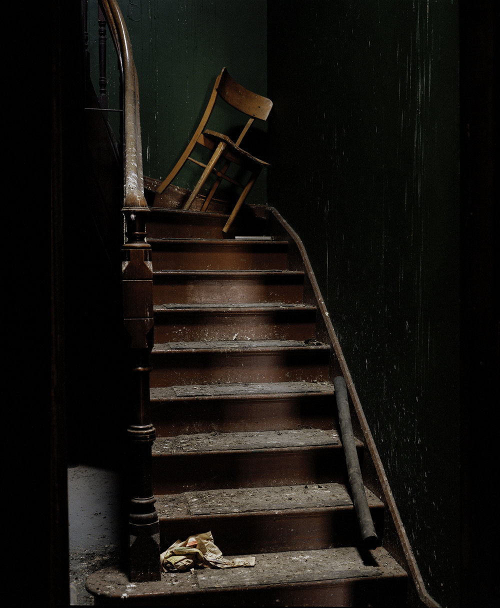 After Magdalene by Ethna Rose O'Regan — A wooden chair lies toppled on metal stairs within the abandoned Magdalene Laundry, its rush seat unraveled across the treads.