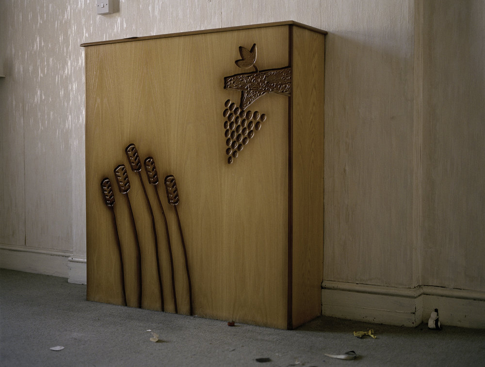 After Magdalene by Ethna Rose O'Regan — A wooden cabinet occupies the corner of a sparse institutional interior, its honey-toned oak surface carved with deeply incised botanical reliefs.