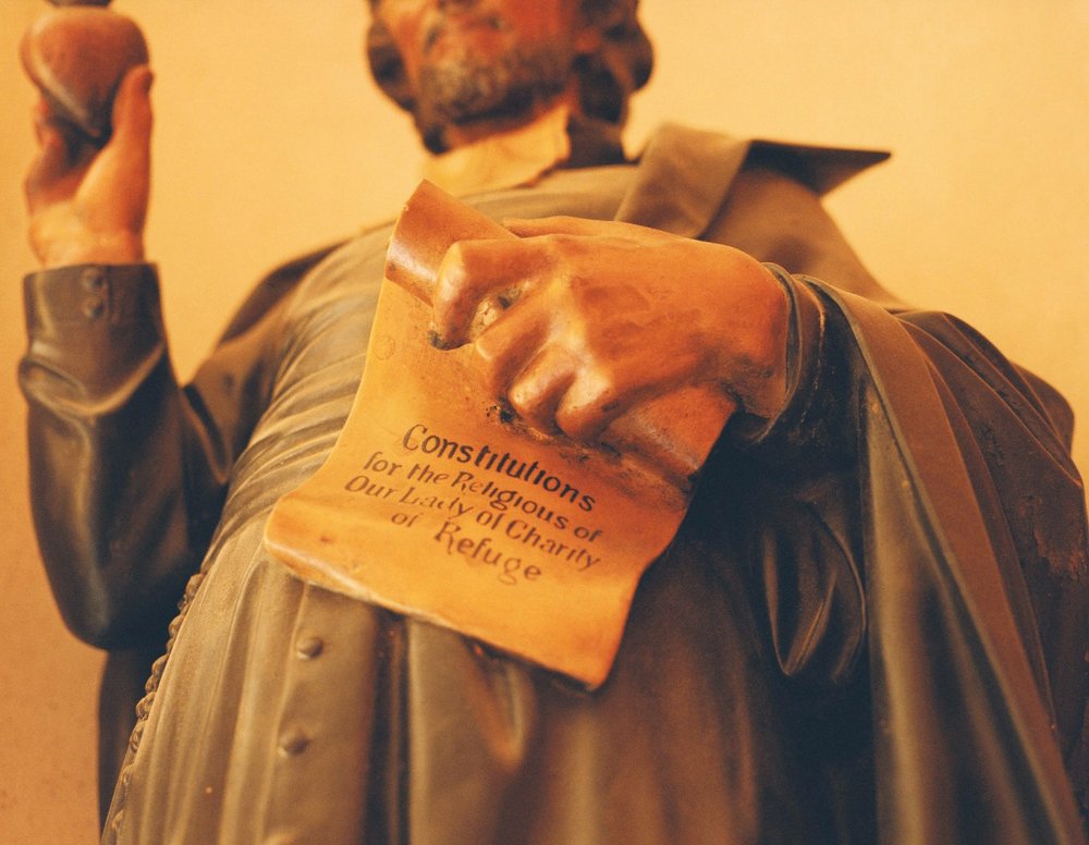 After Magdalene by Ethna Rose O'Regan — A wooden sculptural relief depicts a robed figure holding an inscribed tablet, its surface carved with text reading 'Constitutions for the Religious of Our...