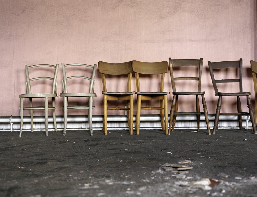 After Magdalene by Ethna Rose O'Regan — Six wooden chairs arranged in a linear row against a pink-rendered wall, their shadows cast sharply across the surface by overhead light.
