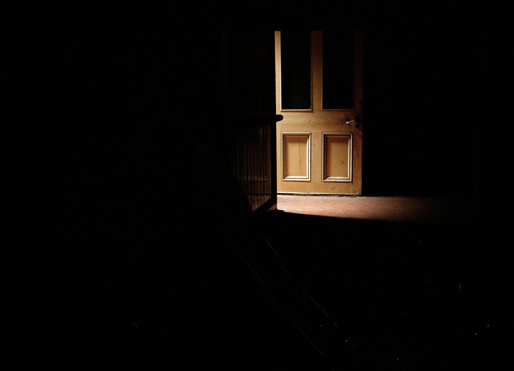 After Magdalene by Ethna Rose O'Regan — A wooden door with two recessed panels dominates the composition, positioned off-center within an arched stone or plaster alcove.