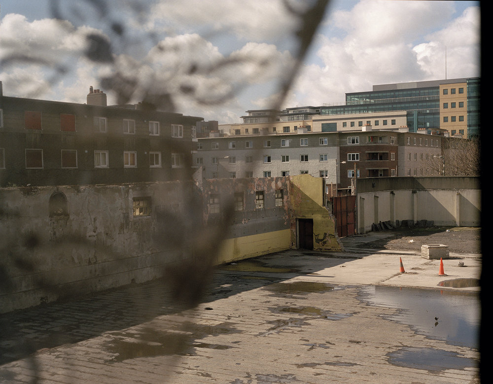 After Magdalene by Ethna Rose O'Regan — A concrete courtyard within the disused Magdalene Laundry on Sean McDermot Street, Dublin, photographed through a wire fence.
