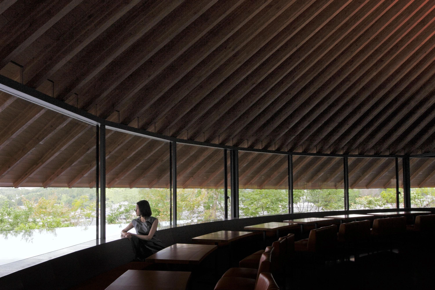 Sayama Lakeside Cemetery Community Hall by Hiroshi Nakamura & NAP — A woman seated on an integrated timber bench surveys the forest canopy through continuous glazing.