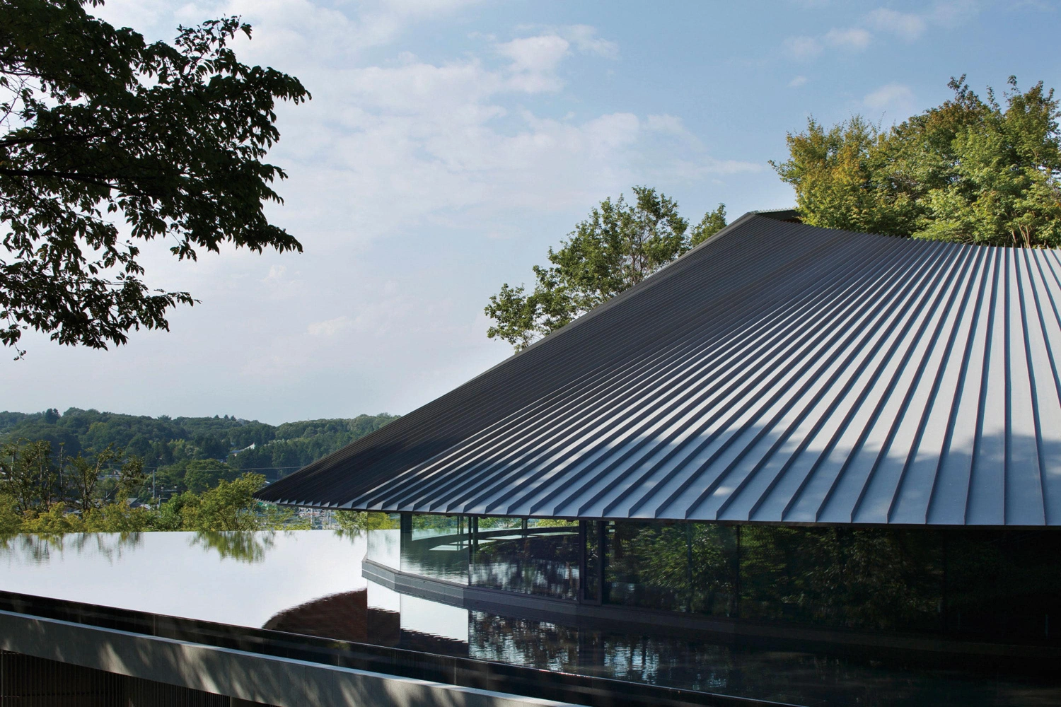 Sayama Lakeside Cemetery Community Hall by Hiroshi Nakamura & NAP — The lakeside elevation reveals the community hall's defining formal gesture: a steeply pitched metal roof with pronounced linear ribbing that descends...