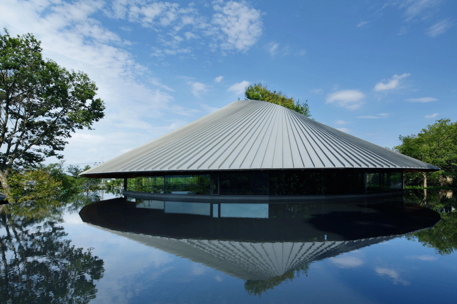 Sayama Lakeside Cemetery Community Hall by Hiroshi Nakamura & NAP — The circular timber-roofed pavilion floats above its reflecting pond, its radial metal roof creating pronounced linear striations that converge toward the apex.