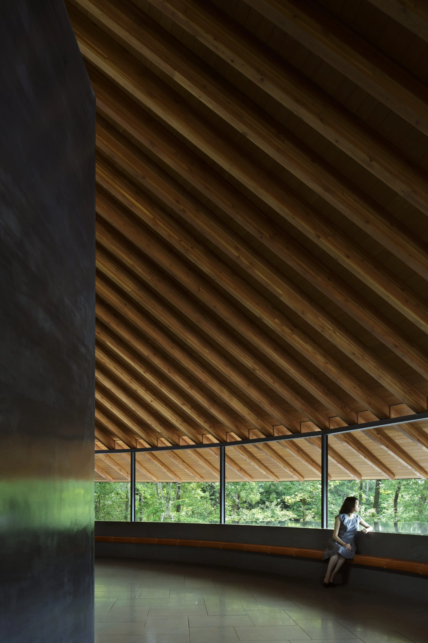 Sayama Lakeside Cemetery Community Hall by Hiroshi Nakamura & NAP — A solitary figure sits on the integrated timber bench positioned along the continuous glazing, gazing toward the forest canopy beyond.