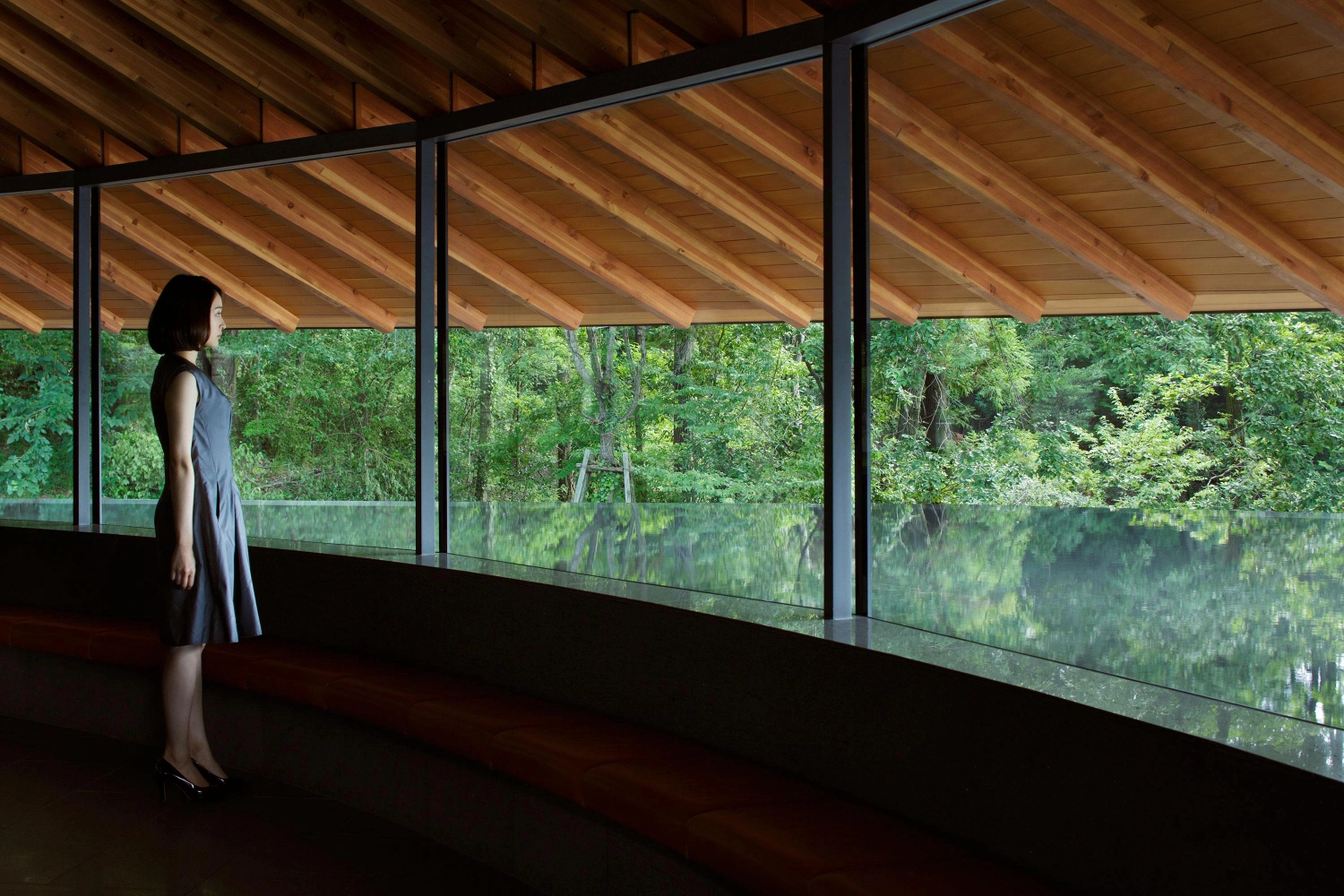 Sayama Lakeside Cemetery Community Hall by Hiroshi Nakamura & NAP — A figure in a sleeveless grey dress stands within the glazed pavilion, positioned at the threshold between interior and forest.