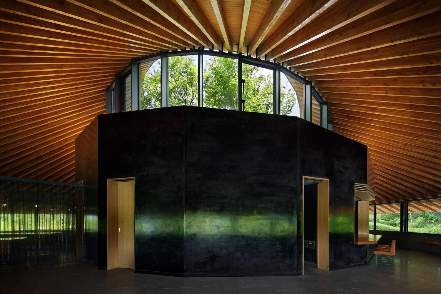 Sayama Lakeside Cemetery Community Hall by Hiroshi Nakamura & NAP — A curved black concrete monolith occupies the center of the timber-framed hall, its weathered surface catching diffused light from the continuous band of...