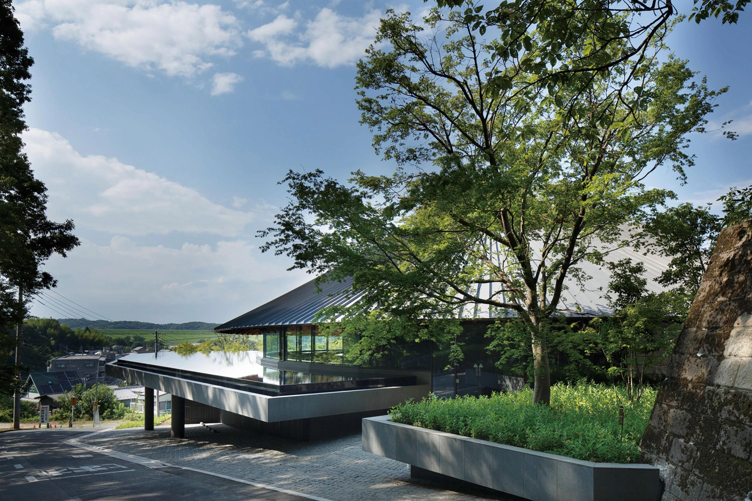 Sayama Lakeside Cemetery Community Hall by Hiroshi Nakamura & NAP — The lakeside community hall emerges from dense woodland, its dark steel-framed structure anchored by a monolithic concrete plinth that rises sharply from...
