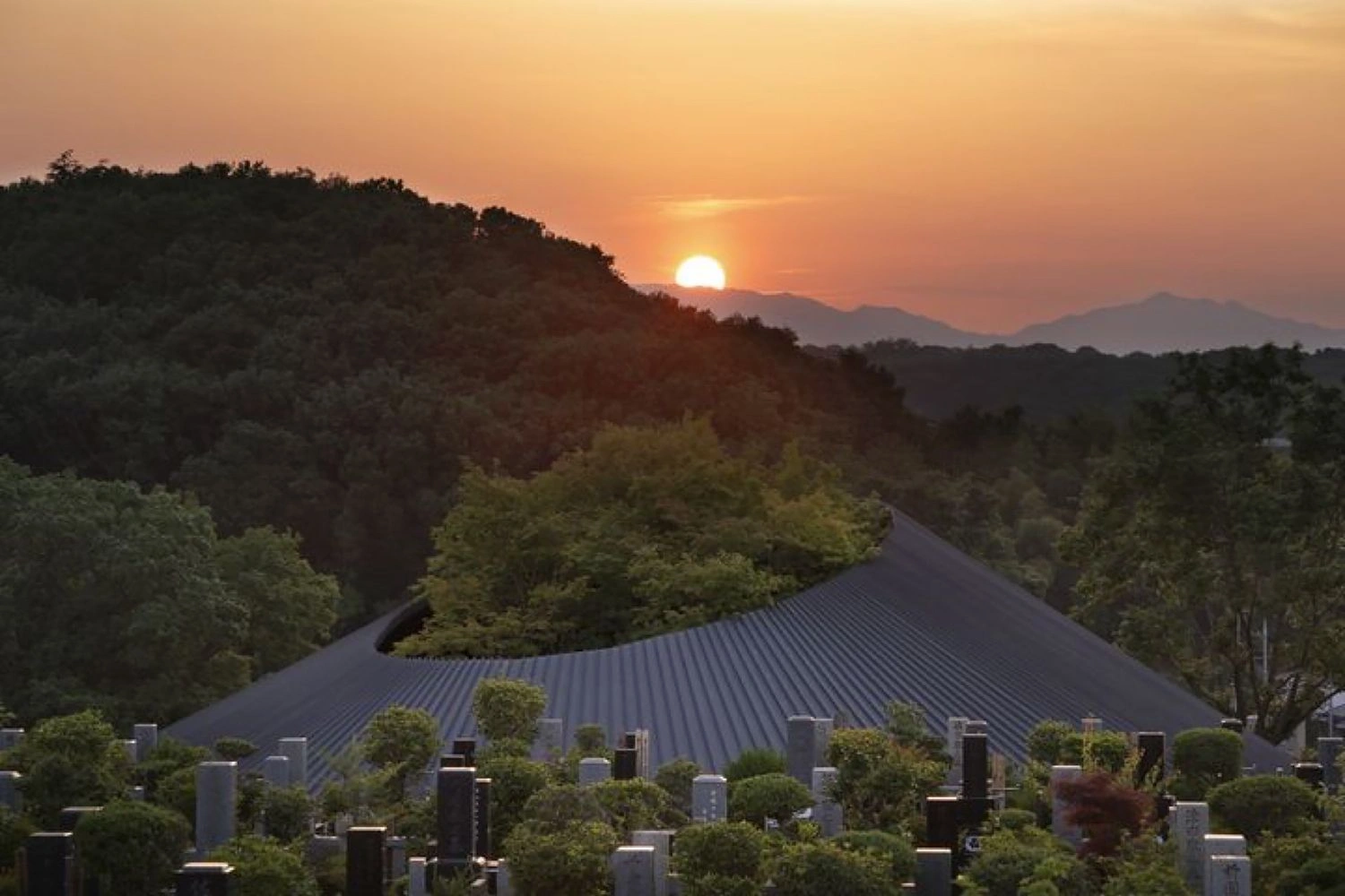 Sayama Lakeside Cemetery Community Hall by Hiroshi Nakamura & NAP — Sunset view across the Sayama Lakeside Cemetery Community Hall reveals the pitched-roof structure's dark metal cladding set against forested hills and...