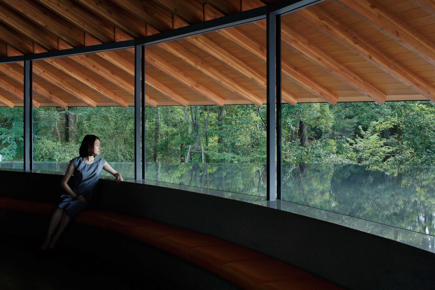 Sayama Lakeside Cemetery Community Hall by Hiroshi Nakamura & NAP — A figure seated on an integrated timber bench gazes across a dark concrete sill toward the forest beyond.