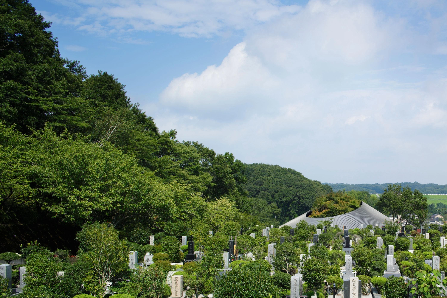 Sayama Lakeside Cemetery Community Hall by Hiroshi Nakamura & NAP — A cemetery landscape dense with white stone monuments and cultivated shrubs, anchored by a pitched-roof pavilion visible in the middle distance.