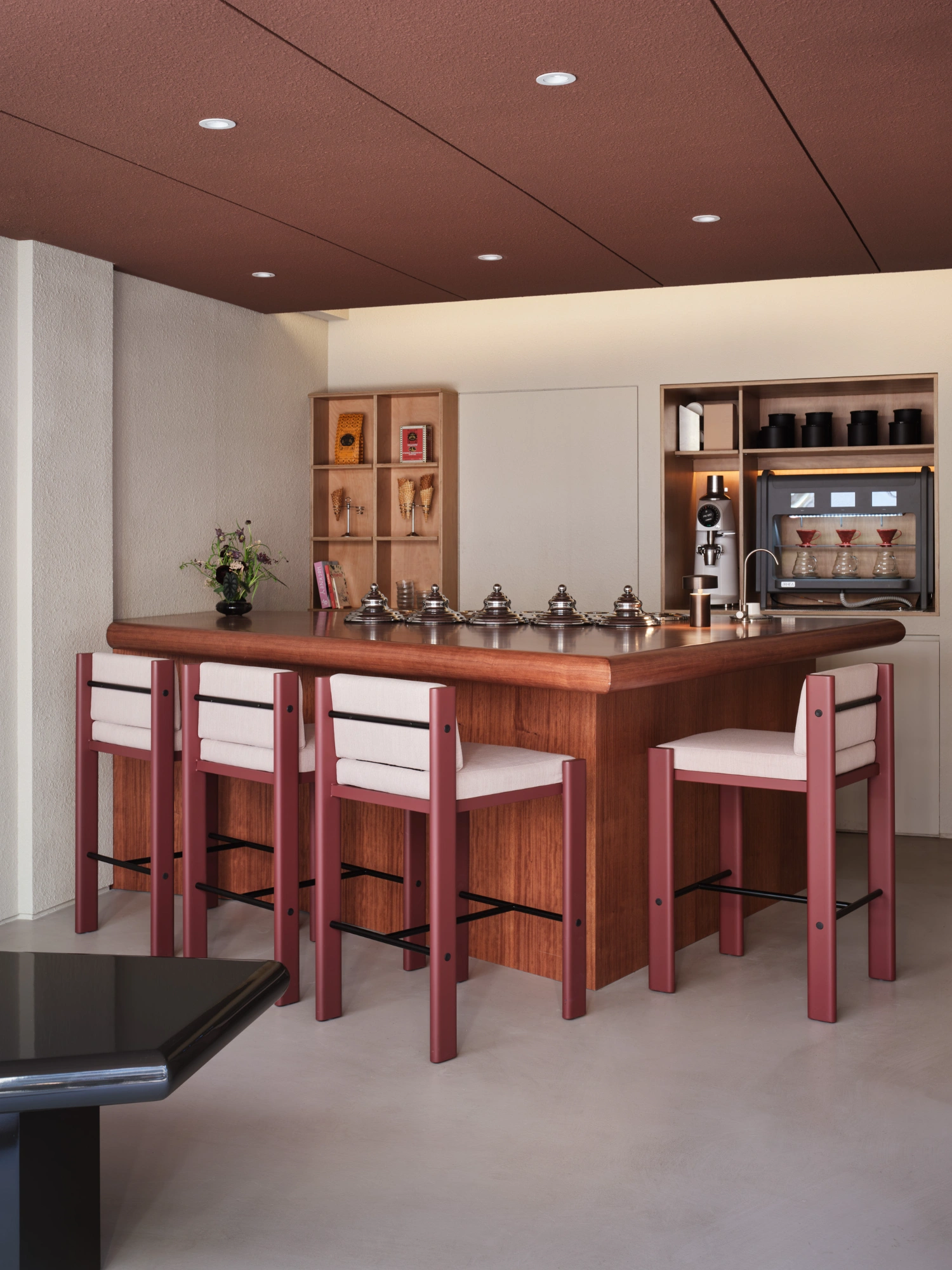 PINOCCHIO Gelato + Coffee Shop by WARP AND WOOF — A bespoke walnut counter with rounded edges anchors the central service area, flanked by four burgundy-painted wooden bar stools with cream upholstered...
