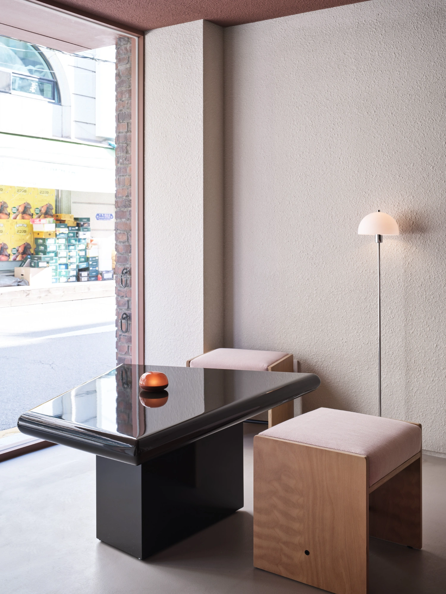 PINOCCHIO Gelato + Coffee Shop by WARP AND WOOF — A minimalist interior moment captured at threshold, where a black-topped desk with rounded edges anchors the foreground beside two natural wood cube stools...