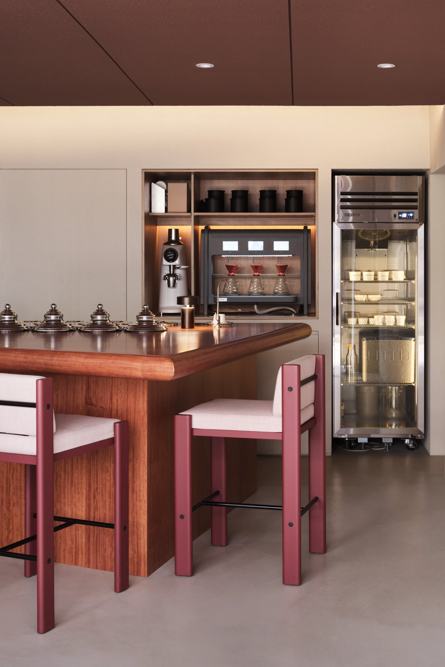 PINOCCHIO Gelato + Coffee Shop by WARP AND WOOF — Interior view of the gelato and coffee counter featuring a walnut-toned wooden serving bar with rounded edges, paired with burgundy-painted wooden stools...