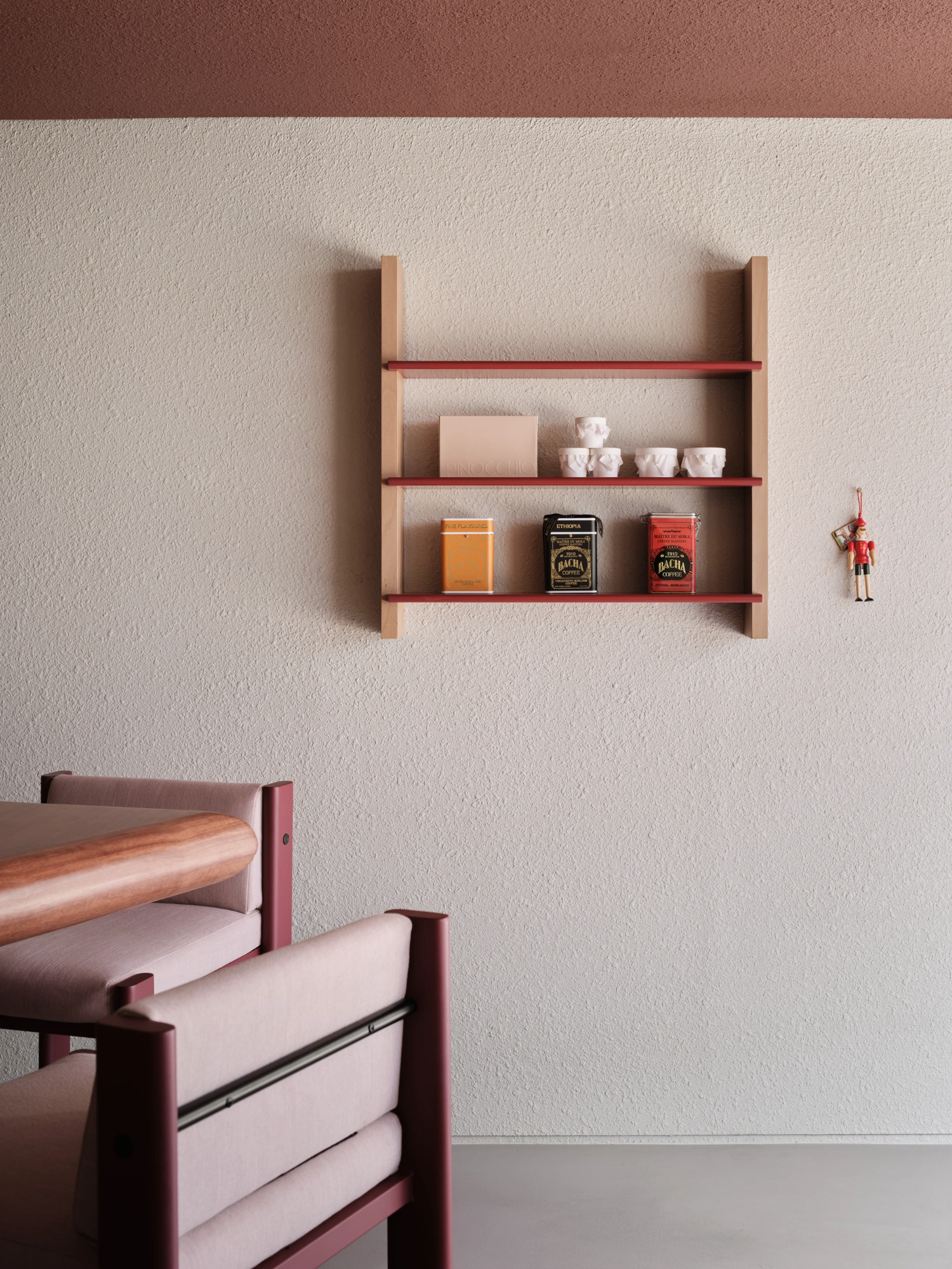 PINOCCHIO Gelato + Coffee Shop by WARP AND WOOF — A wall-mounted shelving unit with natural timber uprights and burgundy metal shelves displays a curated collection of objects: stacked white ceramic...