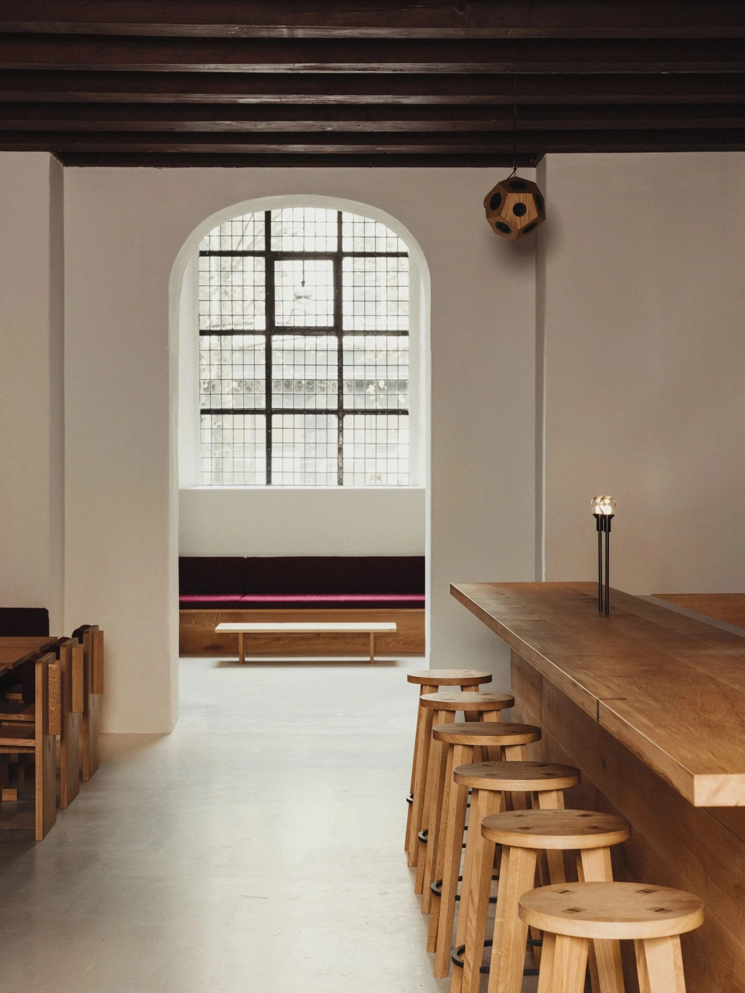 Nikolaj Kunsthal Café + Boutique by Engel Architects by MEE Studio — The café counter occupies the foreground, a long timber bar lined with stacked wooden stools featuring turned legs in natural wood finish.