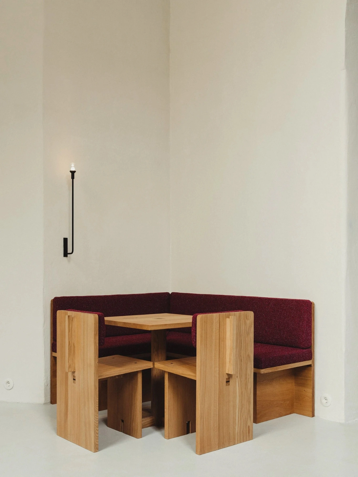 Nikolaj Kunsthal Café + Boutique by Engel Architects by MEE Studio — A compact banquette dining nook with burgundy upholstered seating wraps around a timber table paired with four wooden stools featuring turned legs.