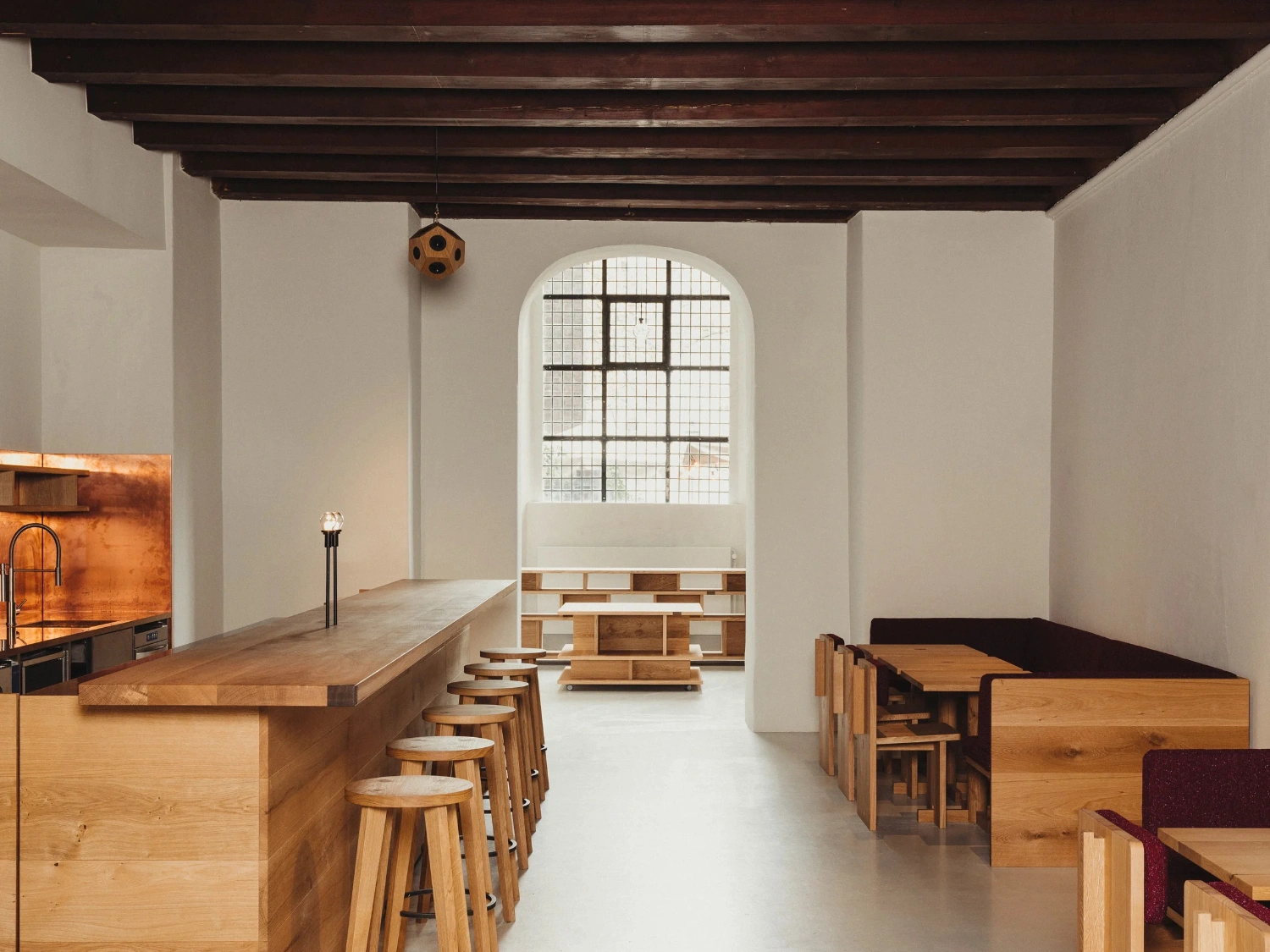 Nikolaj Kunsthal Café + Boutique by Engel Architects by MEE Studio — The café interior showcases a longitudinal spatial organization anchored by a timber bar counter aligned with turned-leg wooden stools in the foreground.