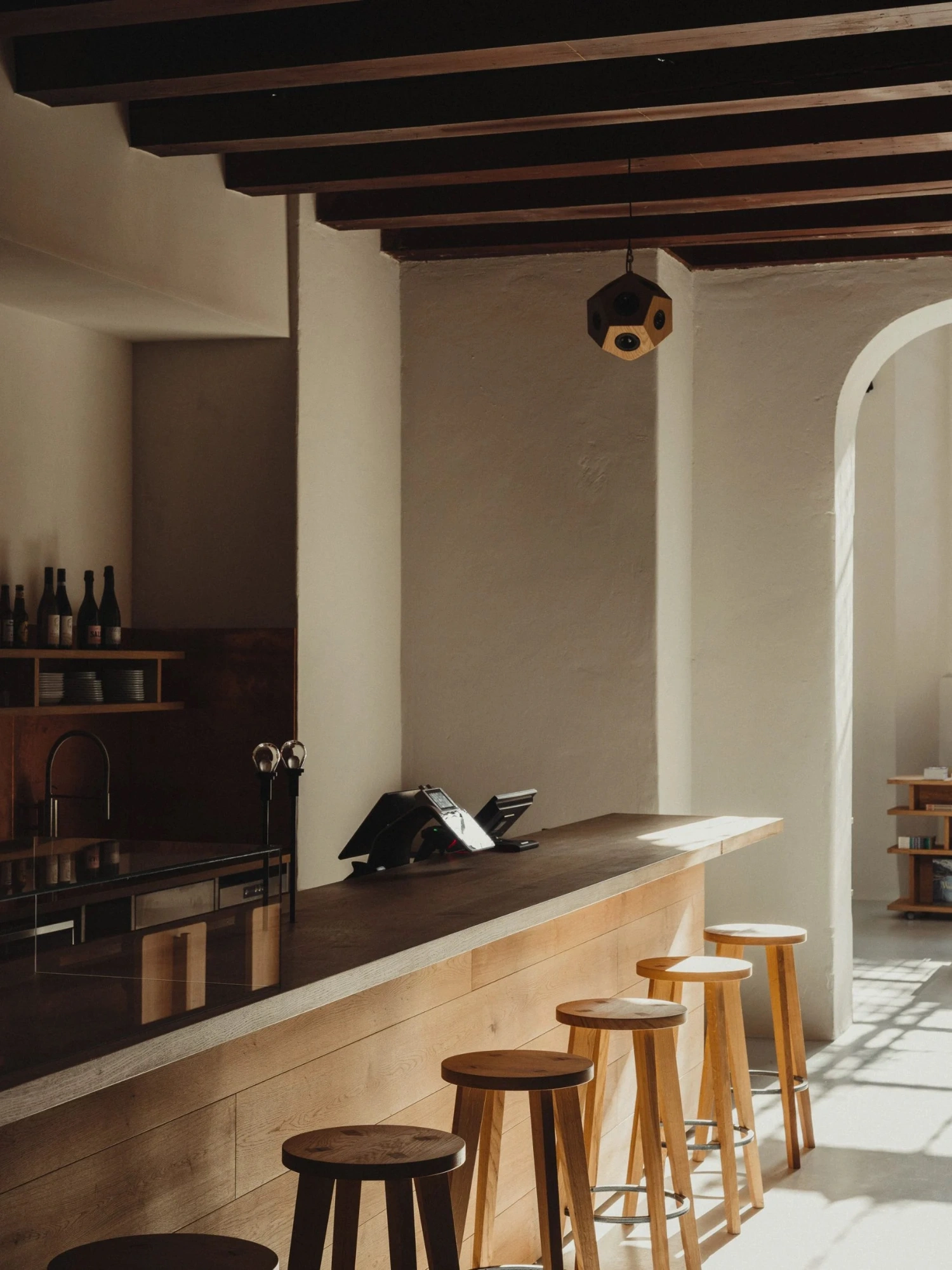 Nikolaj Kunsthal Café + Boutique by Engel Architects by MEE Studio — The café counter anchors the composition with stacked wooden stools featuring turned legs aligned along a timber bar with cream stone top.