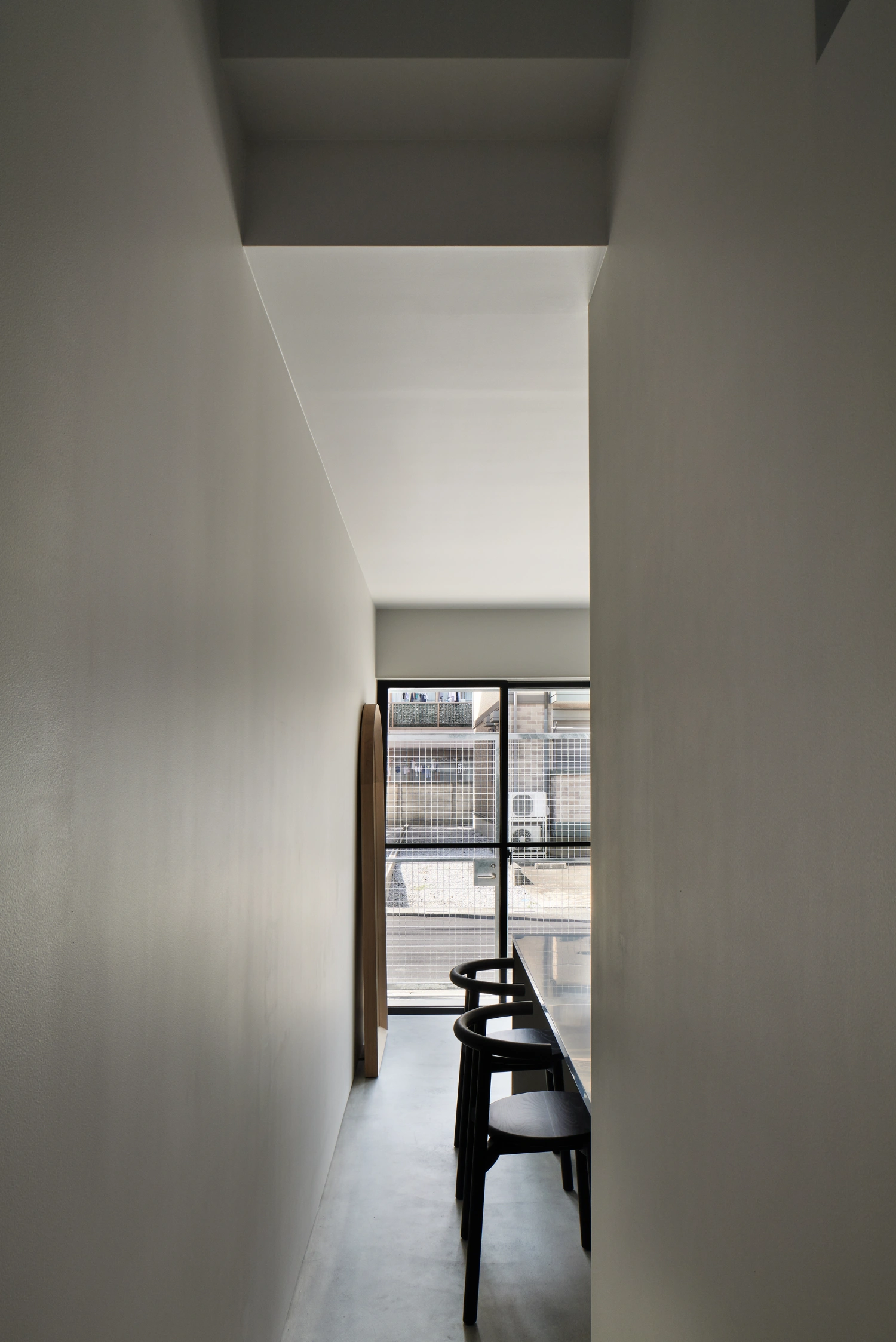 Retreat House by Arii Irie Architects — A narrow corridor frames a black bentwood chair positioned at the threshold between interior and exterior.