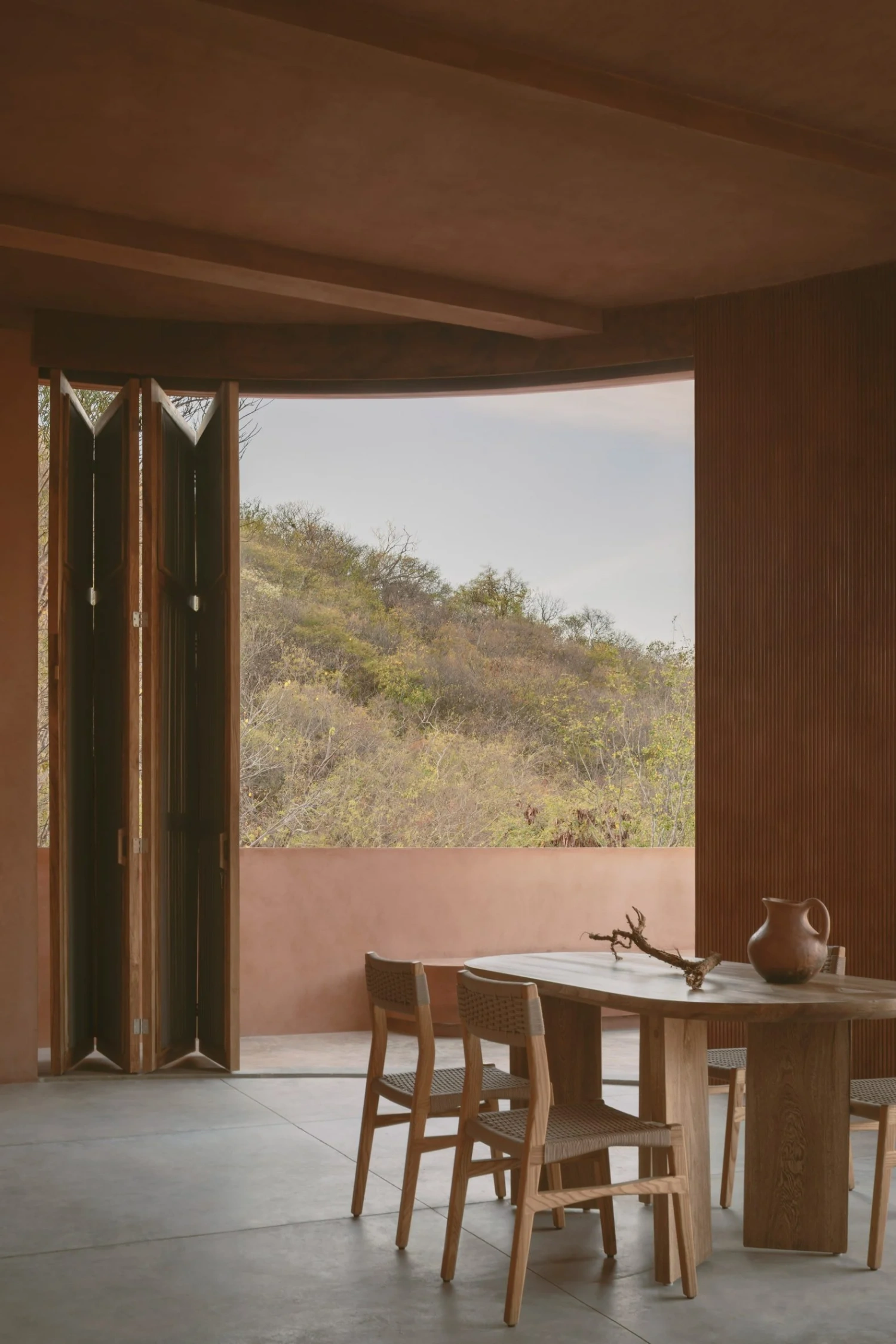 Casa Tobi by Espacio 18 Arquitectura — Bifold wooden doors in warm honey tone frame a threshold to the terrace, their slatted construction casting linear shadows across the polished concrete floor.