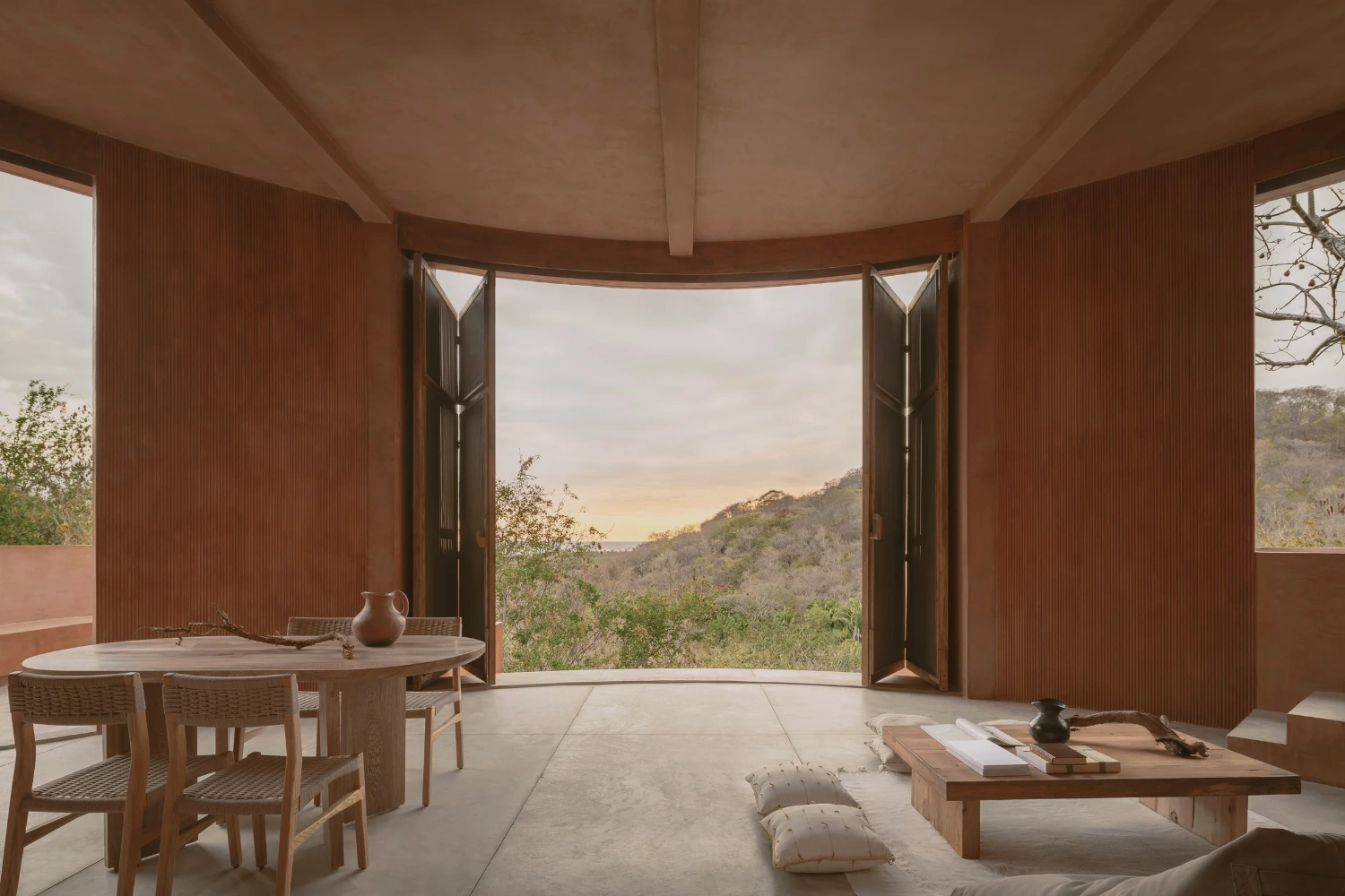 Casa Tobi by Espacio 18 Arquitectura — A cylindrical interior frames landscape through paired timber-slatted doors in honey tone, which open to reveal hillside vegetation and golden-hour sky.
