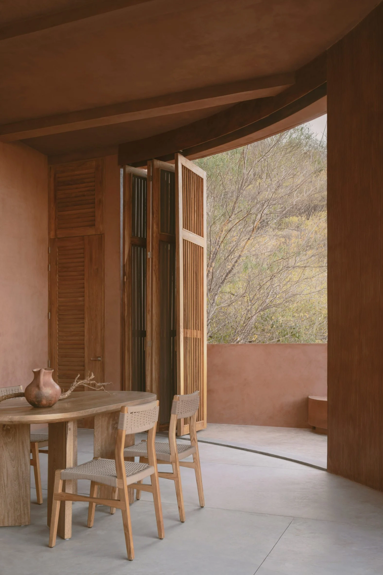 Casa Tobi by Espacio 18 Arquitectura — A threshold moment captured through open wooden slatted doors: interior terracotta render walls frame a dining table in light oak with woven-seat chairs,...