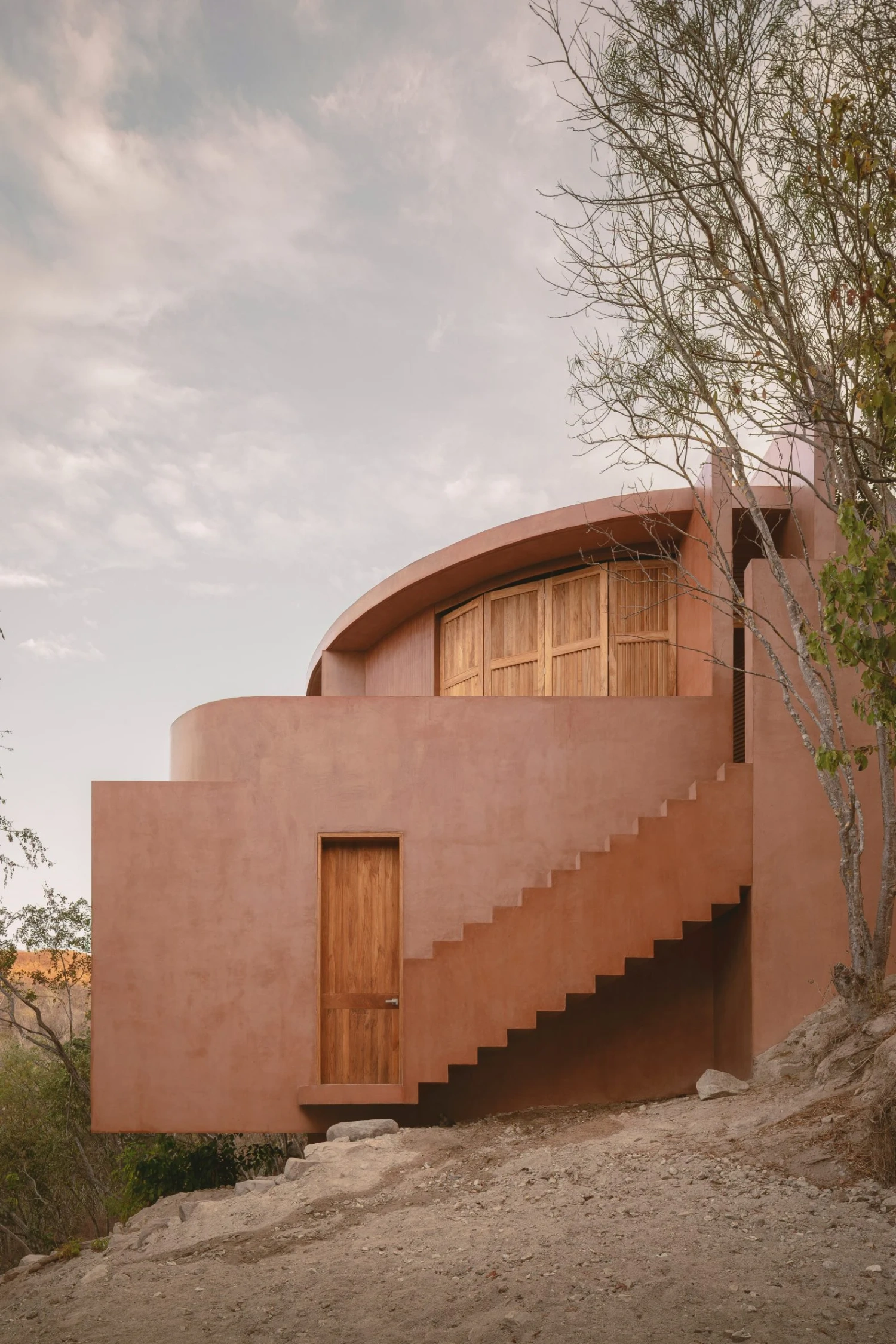 Casa Tobi by Espacio 18 Arquitectura — Casa Tobi's cylindrical volumes in terracotta render descend the sloped terrain via an external stepped staircase, its geometric form anchored by a...