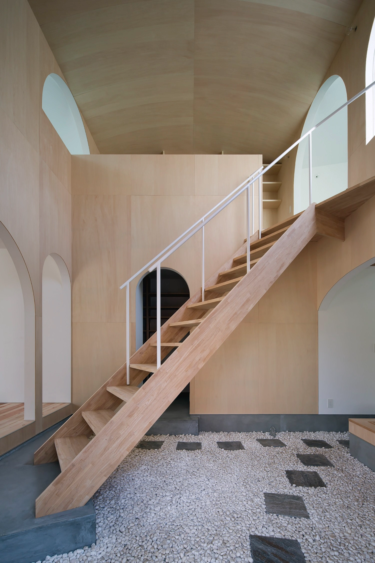 Courtyard in Kudamatsu by Container Design — A diagonal timber staircase traverses the full height of a plywood-lined interior volume, its open treads and white steel railings creating a sculptural...