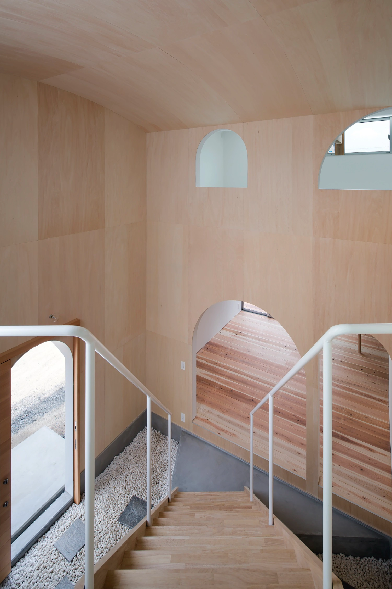 Courtyard in Kudamatsu by Container Design — A diagonal timber staircase with white steel railings ascends through a barrel-vaulted plywood interior, its open treads revealing the layered spatial logic...