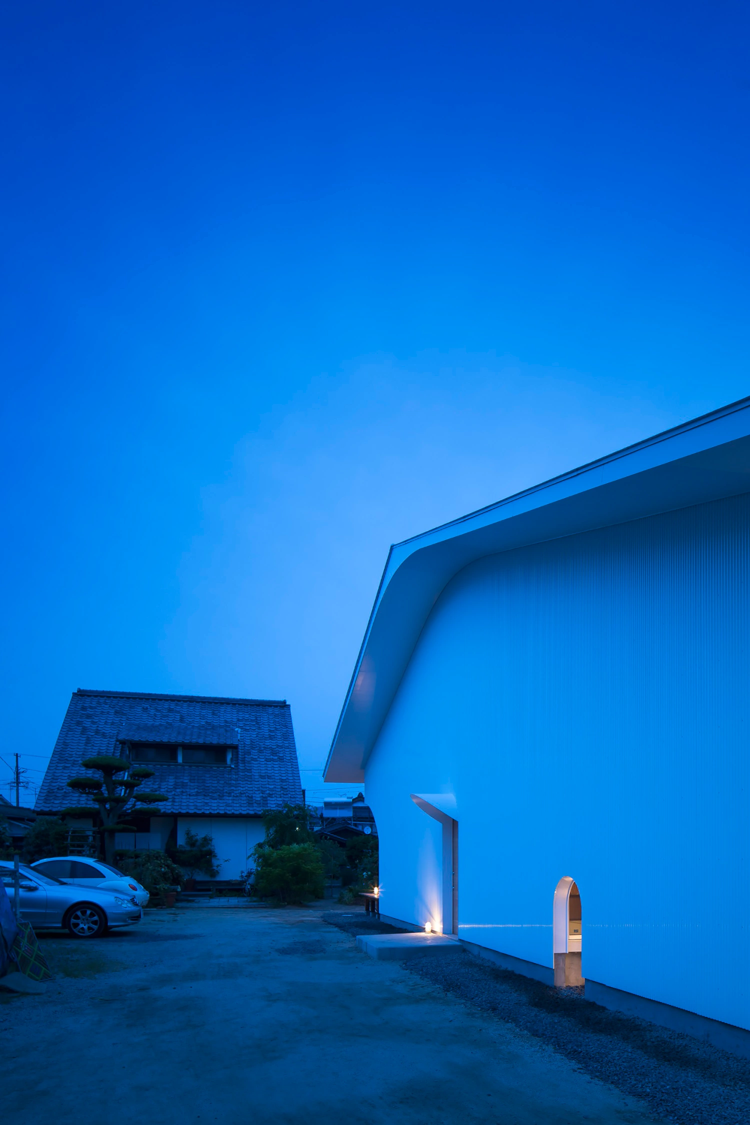 Courtyard in Kudamatsu by Container Design — Twilight view of the courtyard's eastern facade, its curved plywood soffit illuminated against a deep blue sky.