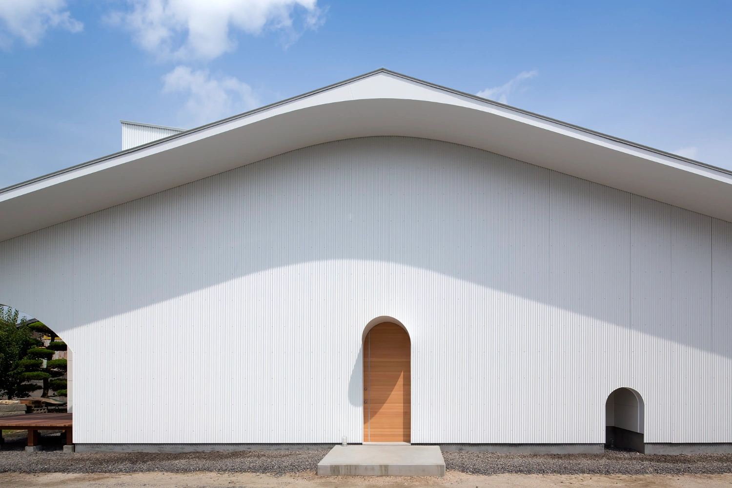 Courtyard in Kudamatsu by Container Design — The barrel-vaulted facade dominates the composition, its white corrugated metal surface cast in sharp shadow beneath the curved roof edge.