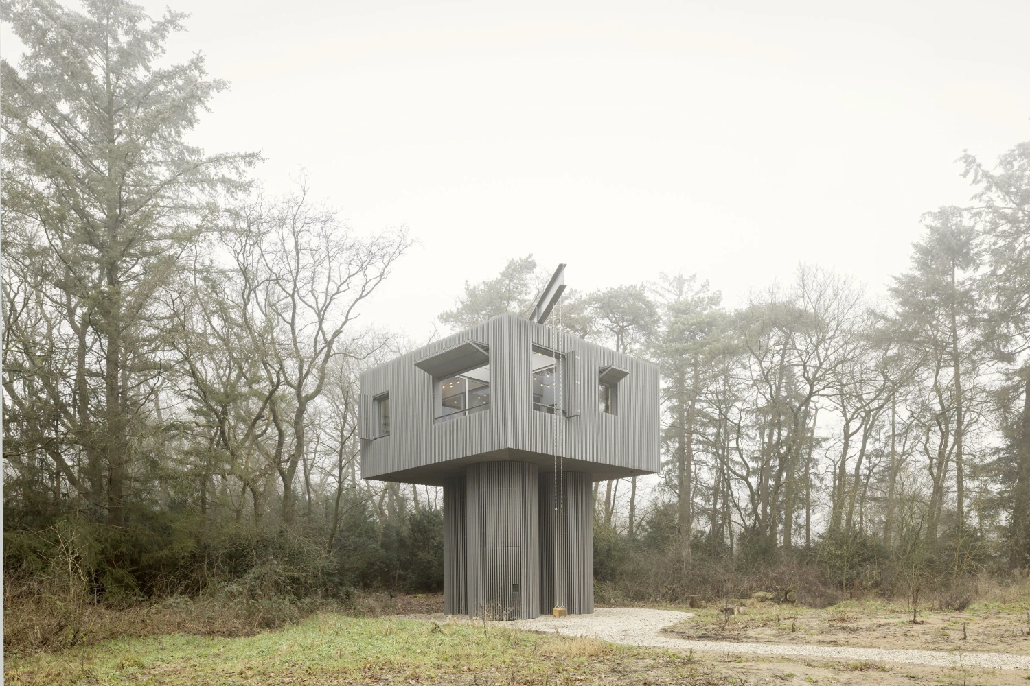 Dolmen by Space Encounters — Space Encounters' Dolmen rises from a forest clearing as a geometric tower, its rectangular upper chamber clad in vertical corrugated metal with uniform...