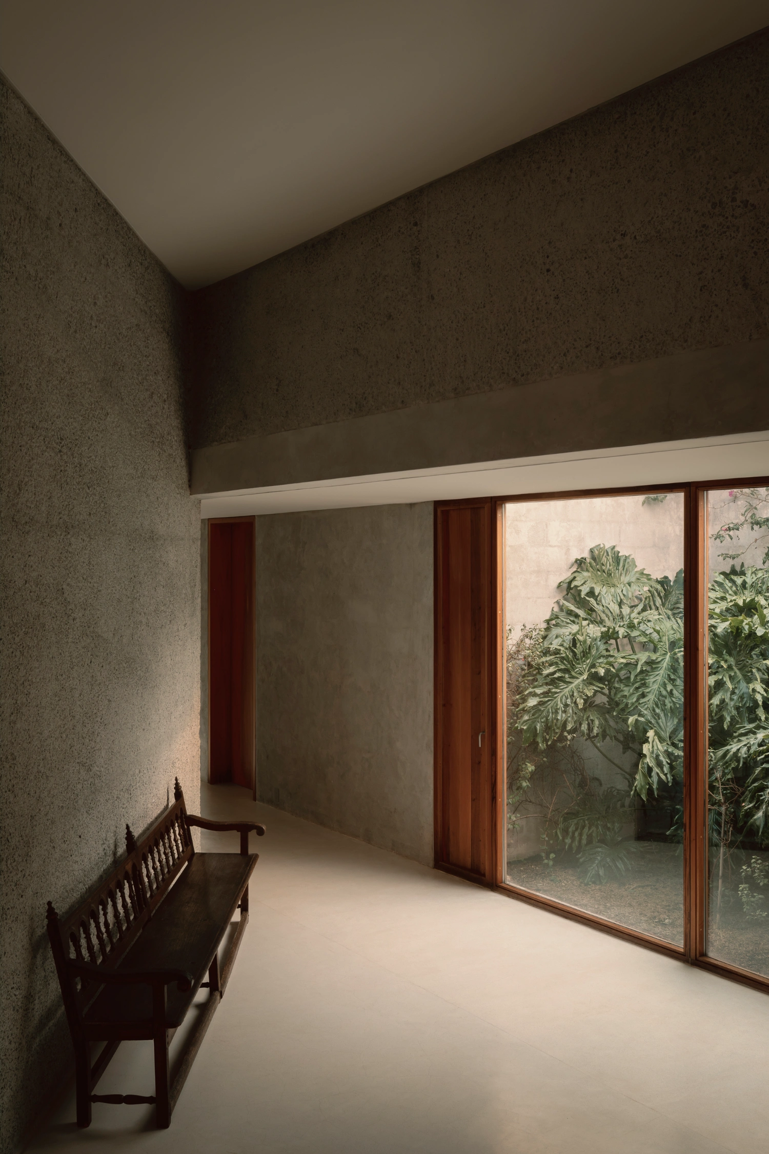 House Between Two Rivers by Taller 3000 — A corridor interior with rendered concrete walls in warm ochre tones transitions toward a threshold framed by timber-mullioned sliding glass doors.