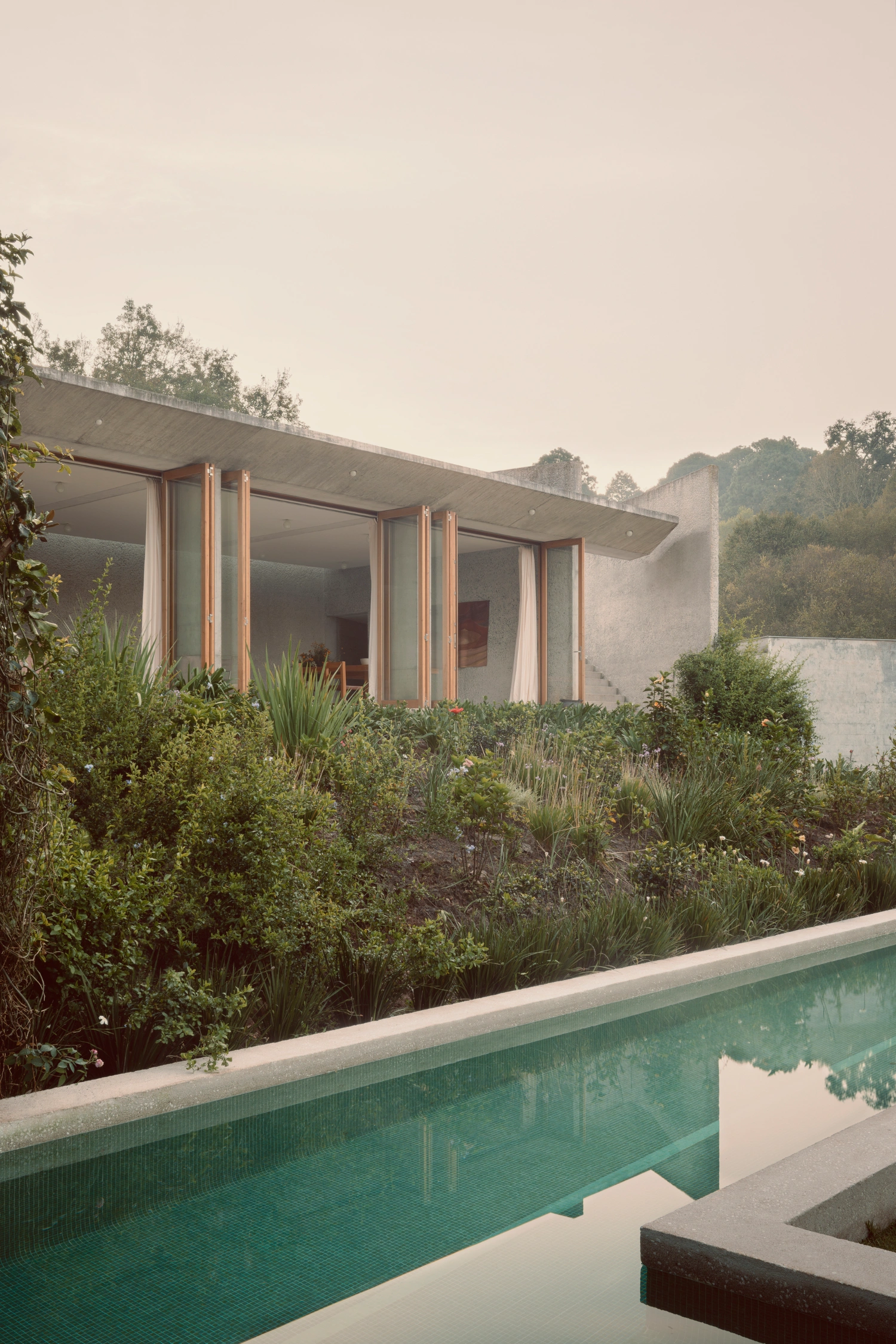 House Between Two Rivers by Taller 3000 — The terrace facade presents a rendered concrete wall punctuated by three tall sliding glass doors with cream-colored curtains, framed by copper or...