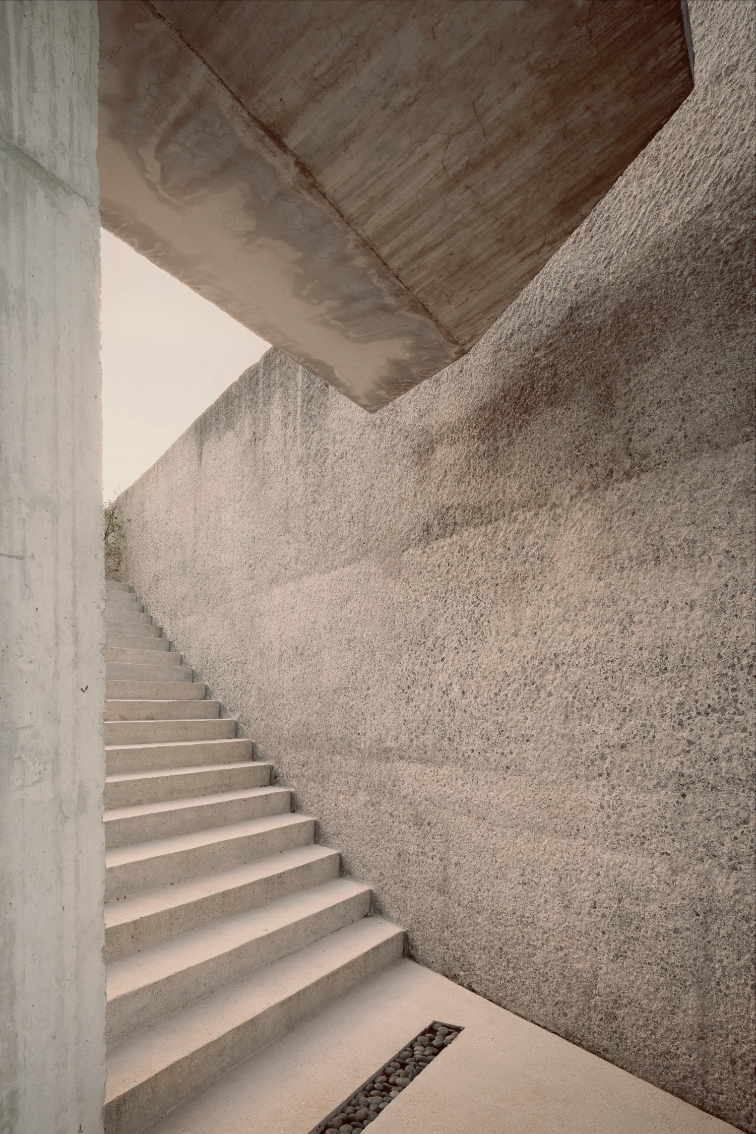 House Between Two Rivers by Taller 3000 — A monolithic concrete staircase descends into the hillside, its trapezoidal form carved against a rendered concrete wall surface marked by weathering and...