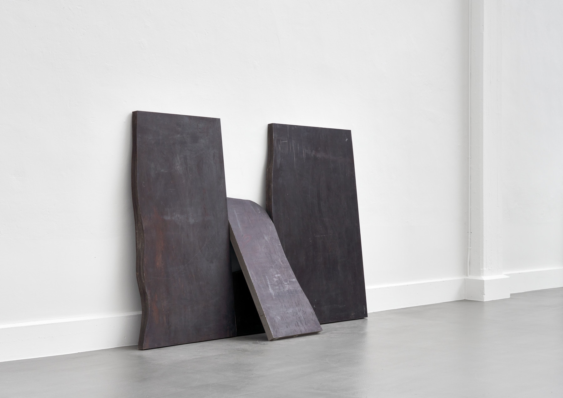Materie Stahl by Alf Lechner at Lechner Museum — Three timber beams in varying states of weathering lean against a white gridded wall in the Lechner Museum gallery.