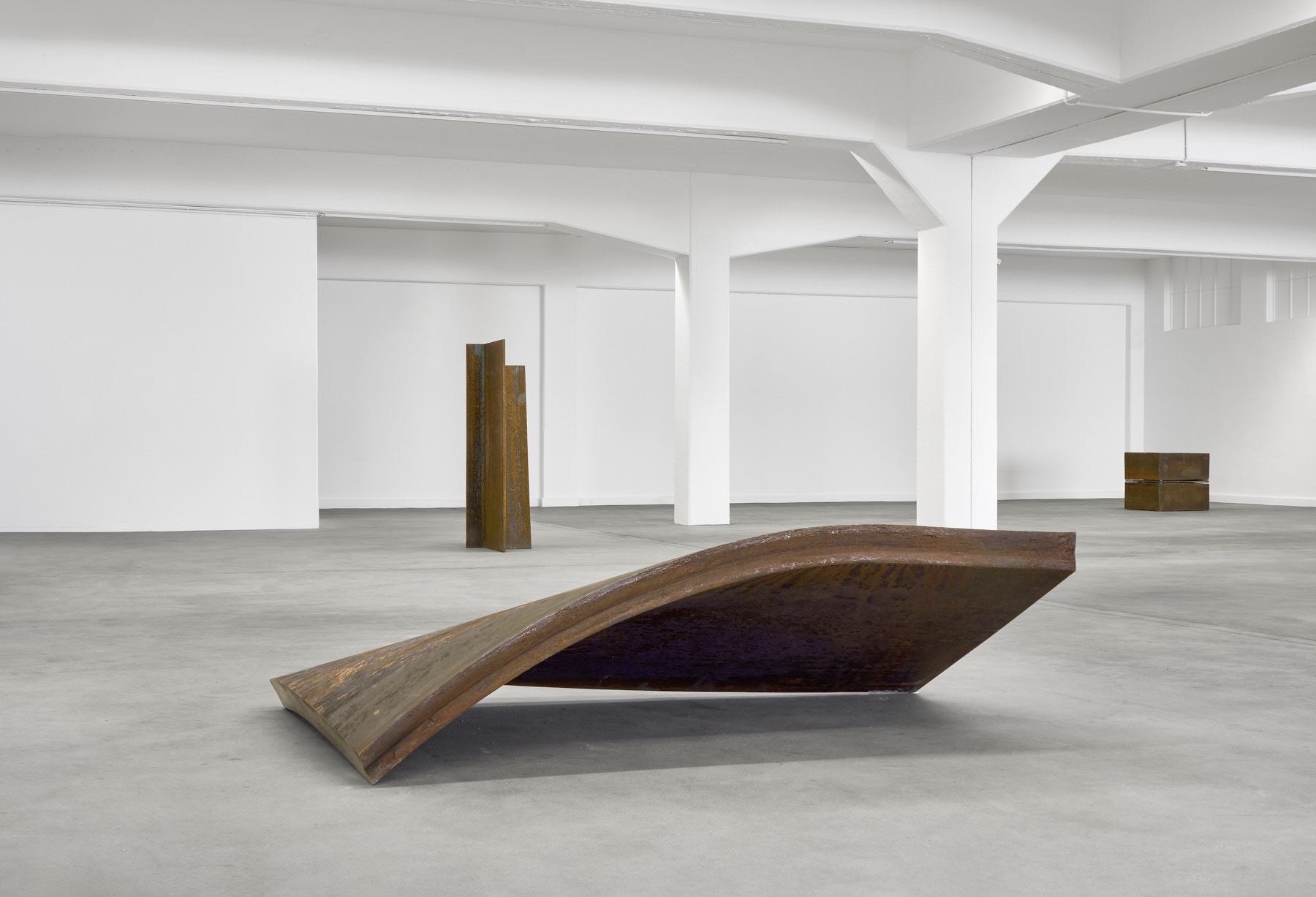 Materie Stahl by Alf Lechner at Lechner Museum — A weathered timber beam, its surface showing deep grain and oxidized patina, tilts at an acute angle across the polished concrete floor.