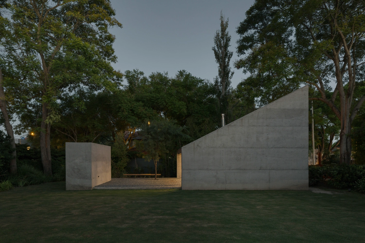 Atelier Workshop by Agustín Berzero — Two monolithic concrete volumes occupy a gravel courtyard at dusk, their raw surfaces catching golden-hour light from a recessed timber-framed glazing strip...